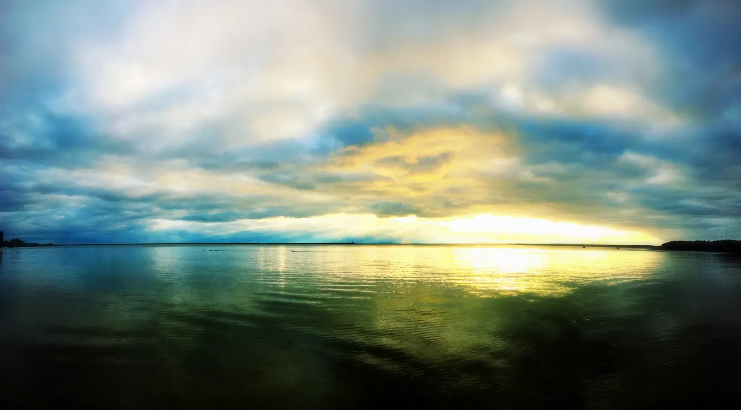 A low sun breaking through cloud over calm open water, its light spread gold across the surface.